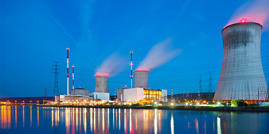 Nuclear power plant with cooling towers and smokestacks reflecting on water at dusk.