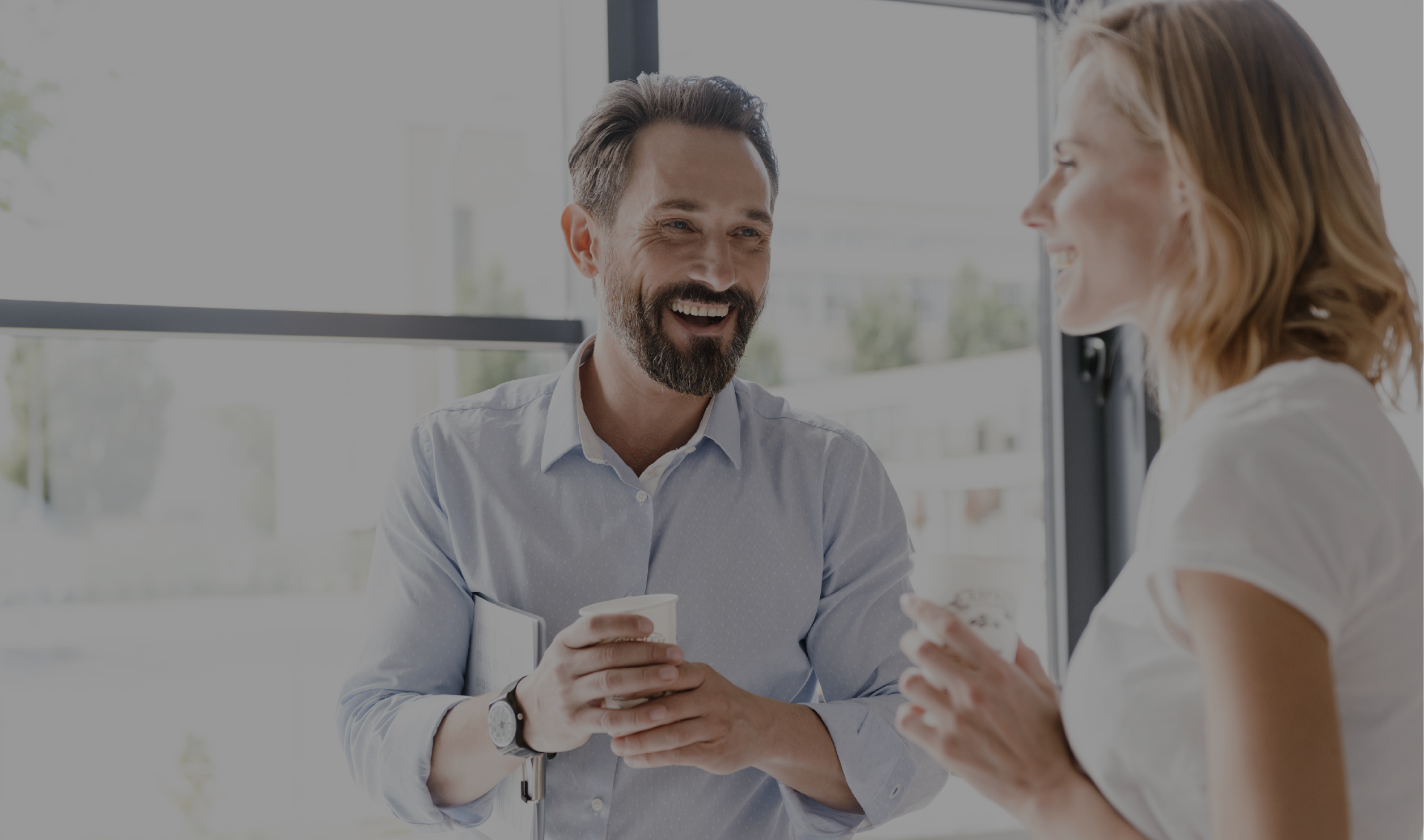 Two professionals smiling and having a casual conversation with coffee in hand, representing a friendly and collaborative business environment.